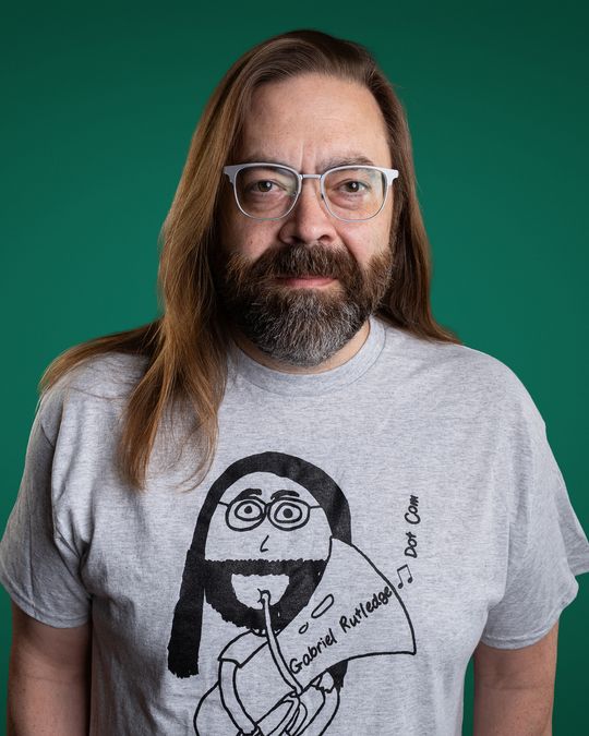 Gabriel Rutledge deadpan in his own cartoon-tuba merch t-shirt, green backdrop